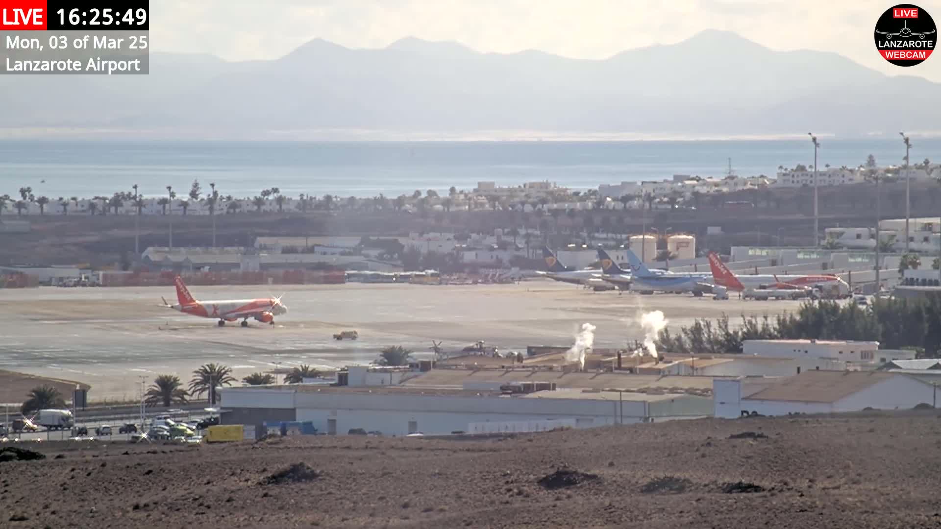 A long shot of an airport tarmac with several airplanes, some buildings, and a calm ocean in the background under a hazy sky.