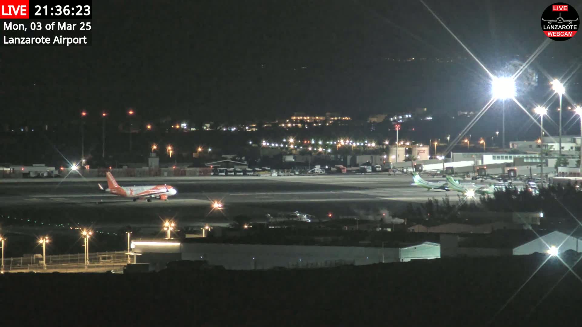 A nighttime view of an airport tarmac with several airplanes, including one orange and white plane taxiing, under clear skies.