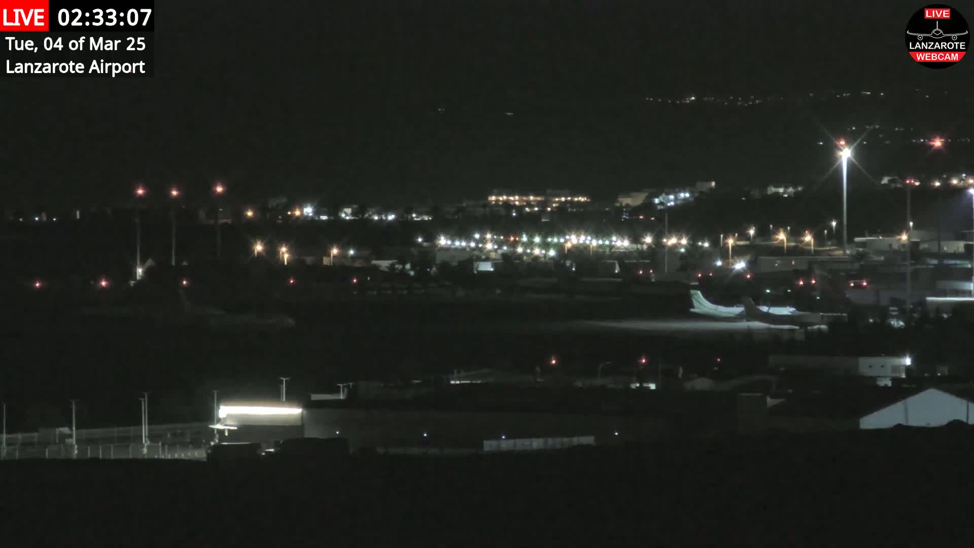 A nighttime view of an airport with numerous lights illuminating the tarmac and surrounding buildings under a clear, dark sky.