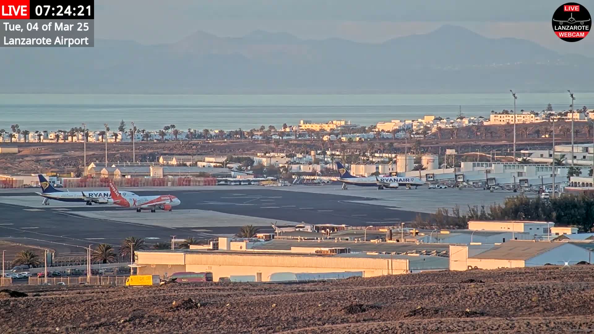Two Ryanair airplanes are parked on an airport tarmac near a body of water and distant mountains under a clear, sunrise sky.