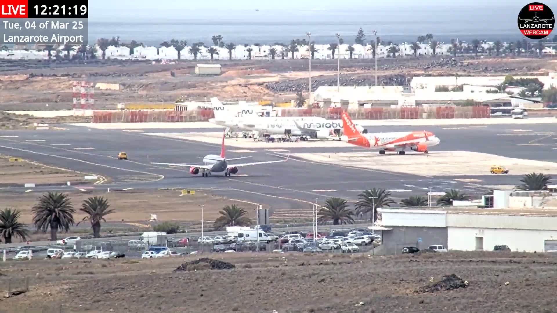 Under a mostly clear sky, several airplanes are parked on an airport tarmac, with cars and buildings visible in the surrounding area.