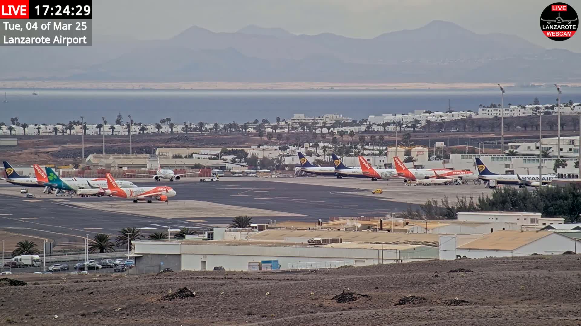 Several airplanes are parked on an airport tarmac near a body of water and mountains under an overcast sky.