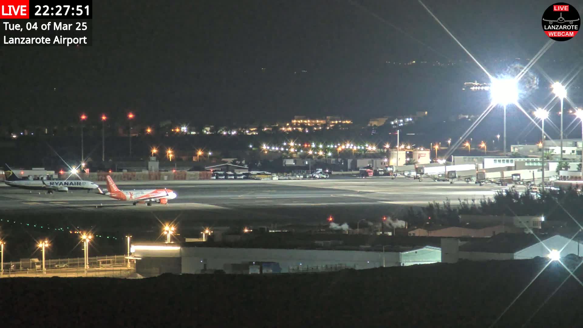 A nighttime view of an airport tarmac with two airplanes, one of which is a Ryanair plane, under clear skies and bright artificial lights.