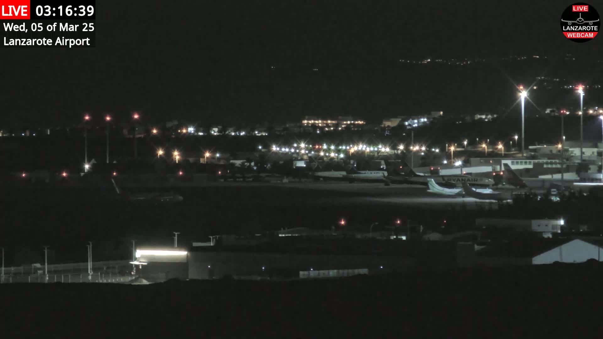 A nighttime long shot of an airport tarmac with several airplanes parked and illuminated by lights, under a clear, dark sky.