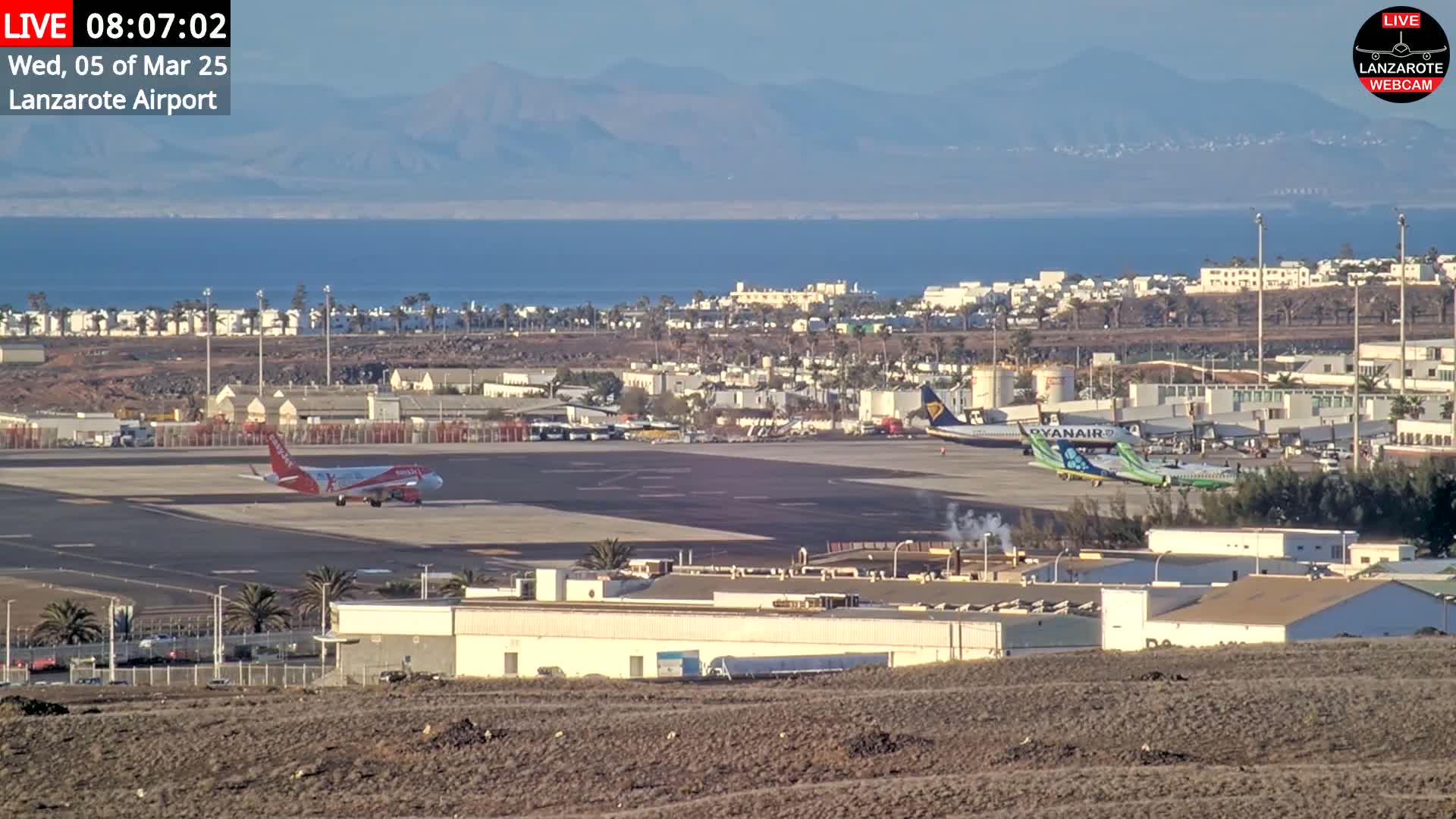 An airport tarmac with several airplanes, including one taxiing, is seen under a clear sky, overlooking a calm ocean and distant mountains.