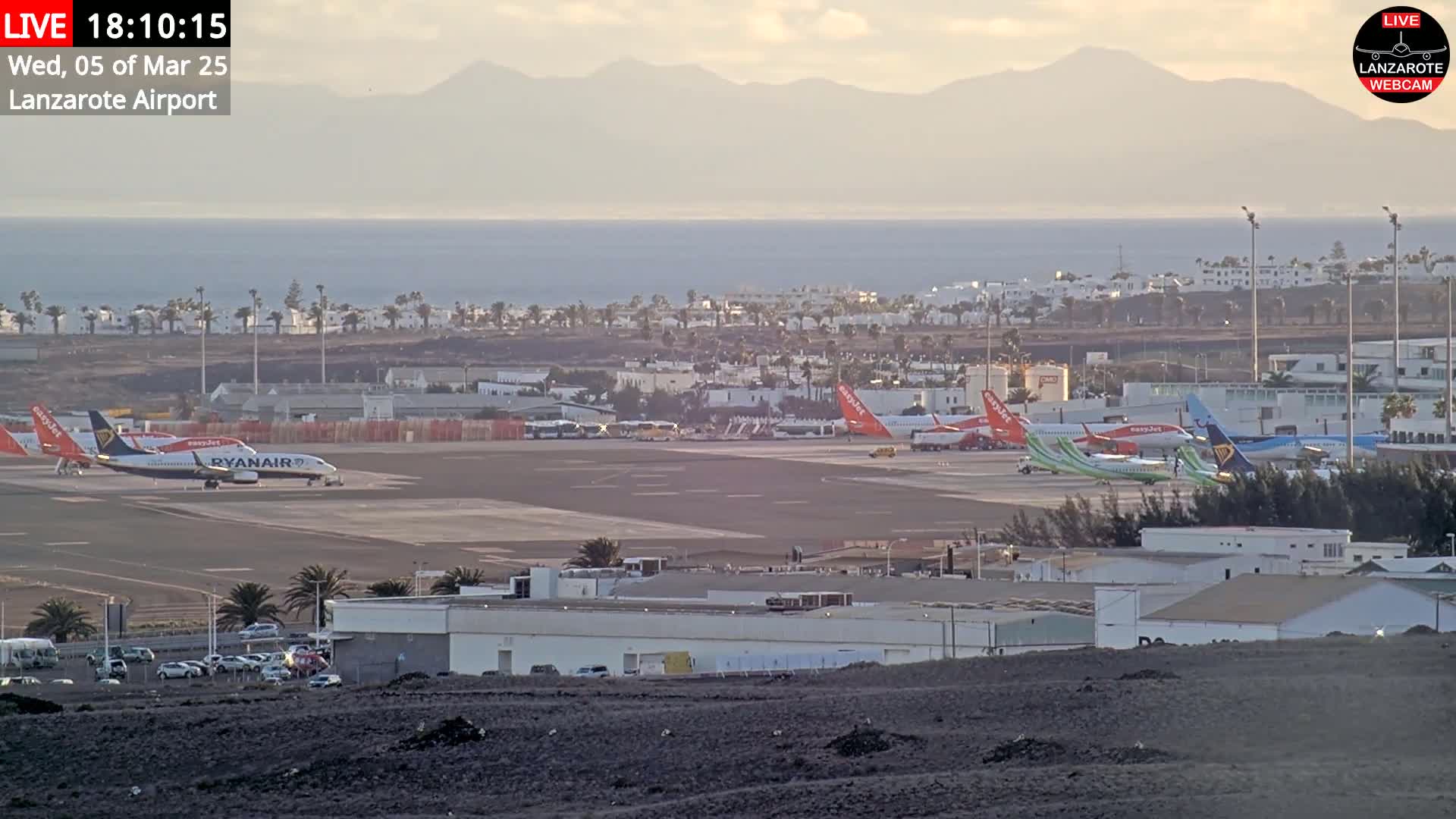 Several airplanes are parked on a tarmac at an airport near a calm ocean and distant mountains under a hazy sky.