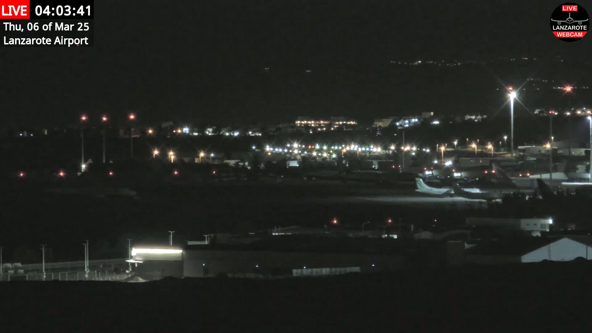 A nighttime, long shot of an airport with several planes on the tarmac, surrounded by dimly lit buildings and structures.