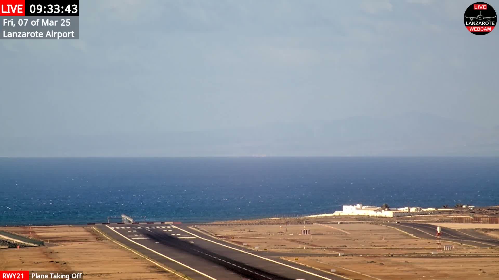 A plane is taking off from an airport runway next to a calm ocean under a mostly clear sky.