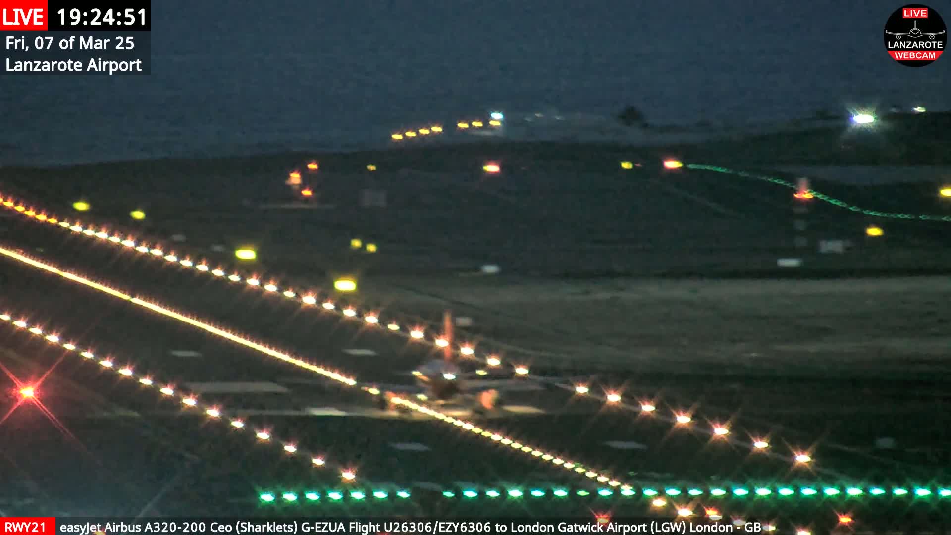 A nighttime view of an airplane taxiing on a runway illuminated by lights, with the ocean visible in the background under a dark sky.
