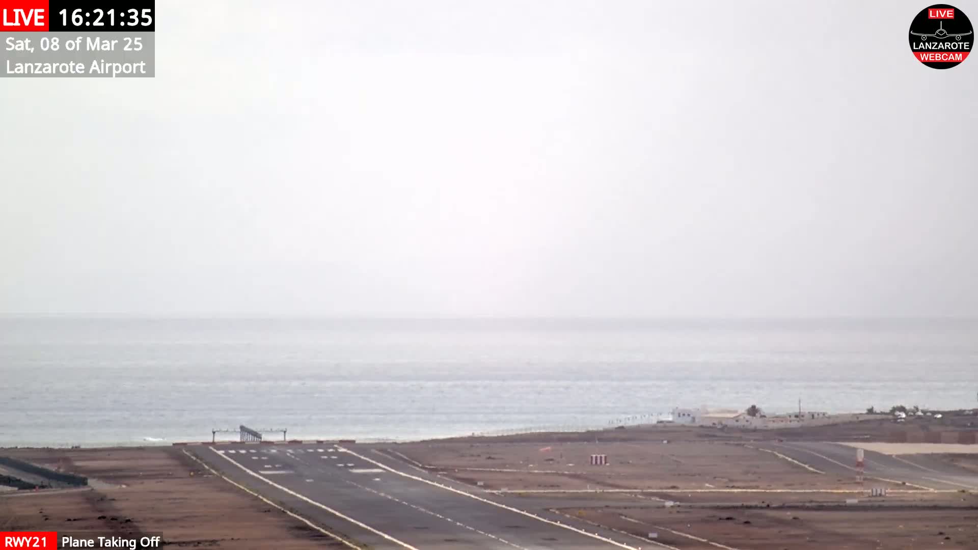 A runway at a coastal airport on an overcast day shows a plane taking off toward a calm ocean.