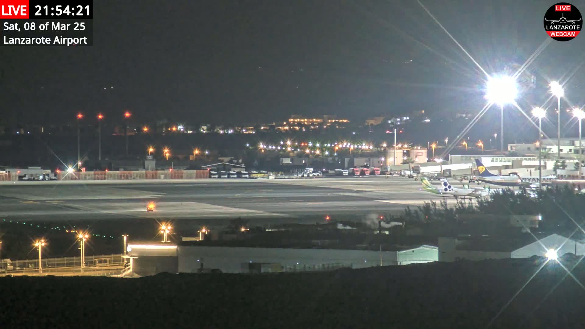 A nighttime view of an airport tarmac with several airplanes and brightly lit buildings in the background under a clear sky.