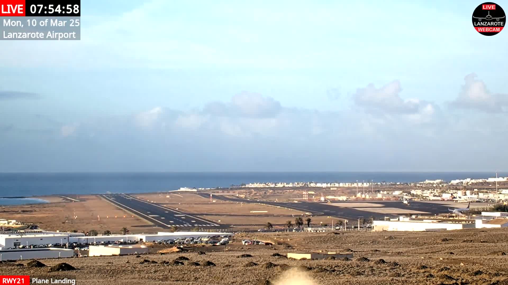 An airport runway and terminal are visible near a calm ocean under a partly cloudy sky.