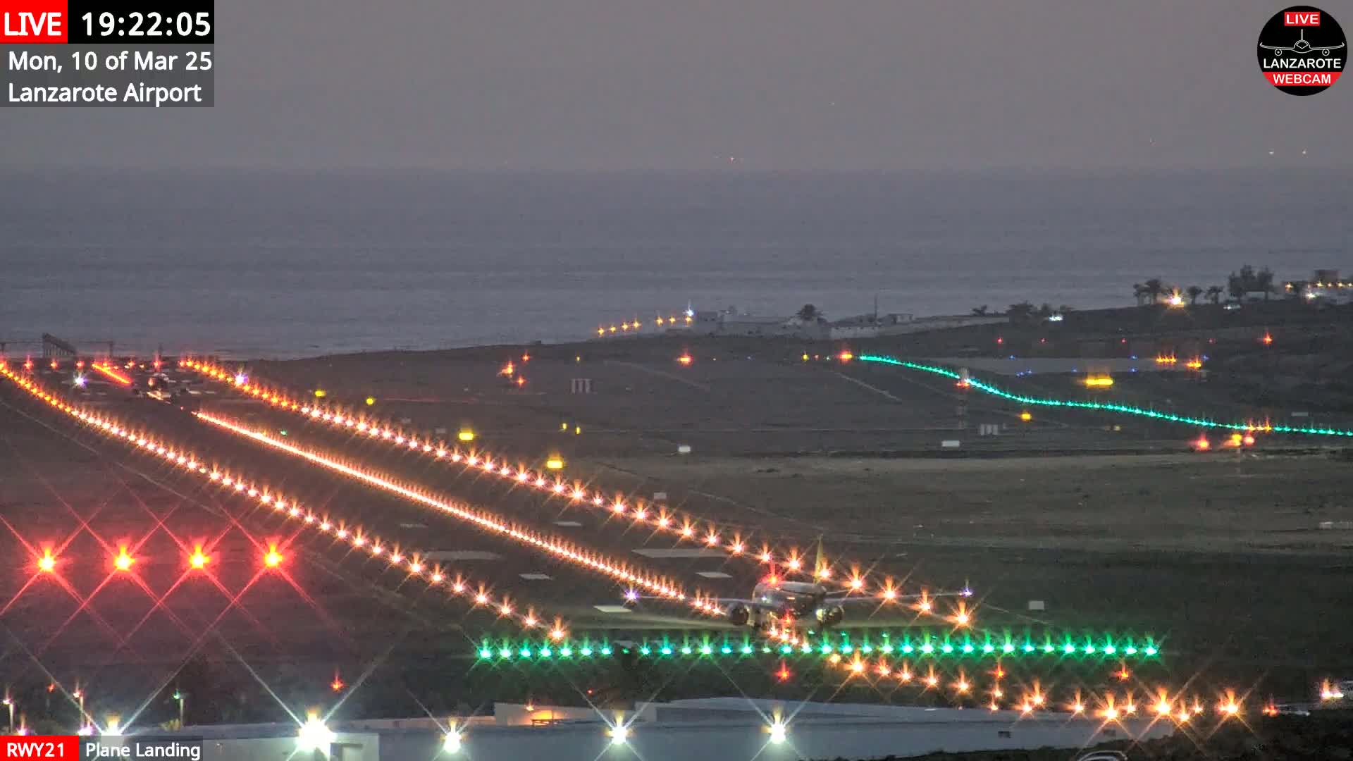 A plane is landing on a brightly lit runway at night, next to a dark ocean under a dusky sky.