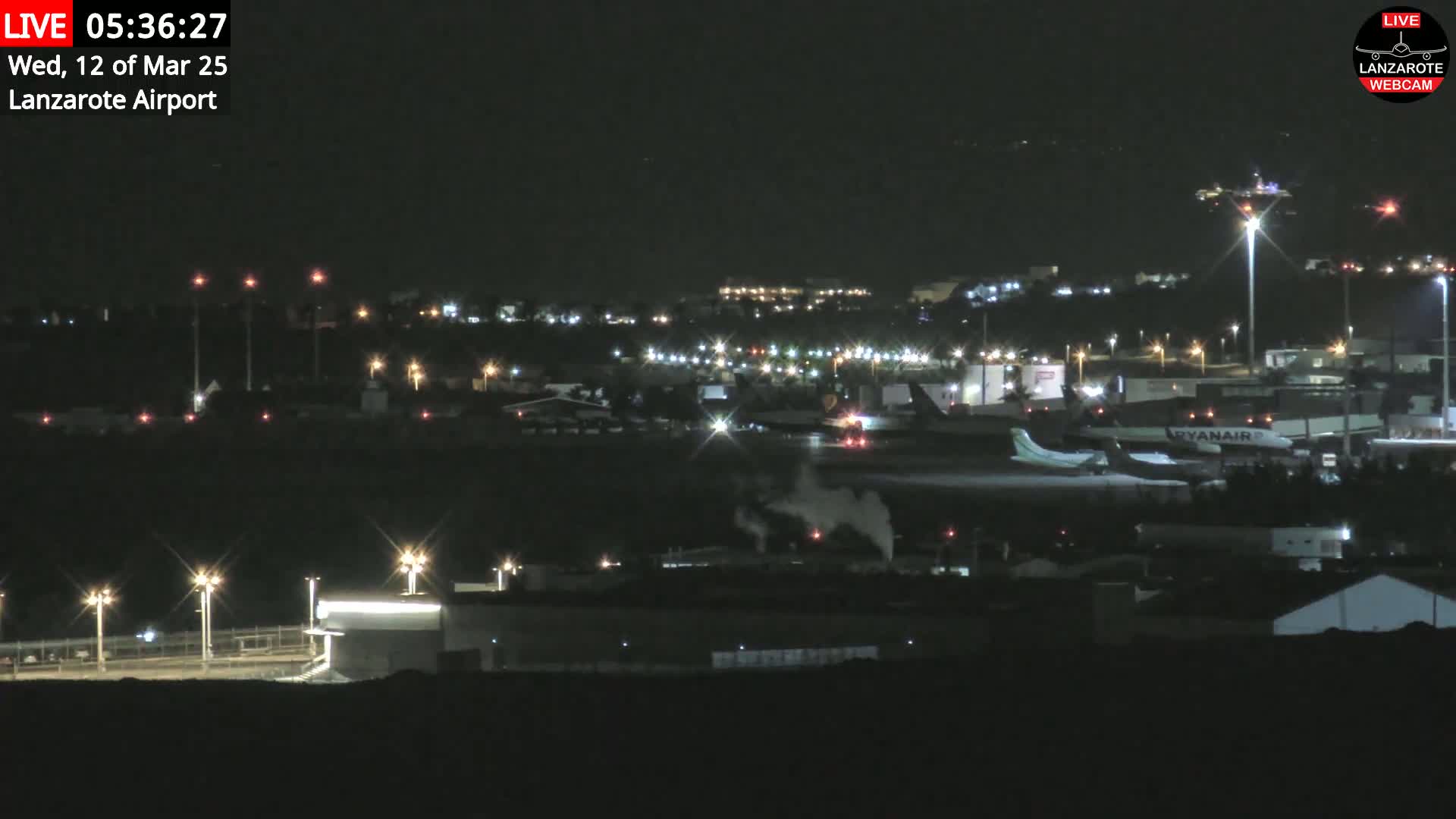 A nighttime view of an airport with several planes on the tarmac, illuminated by runway lights and surrounding building lights under a clear, dark sky.