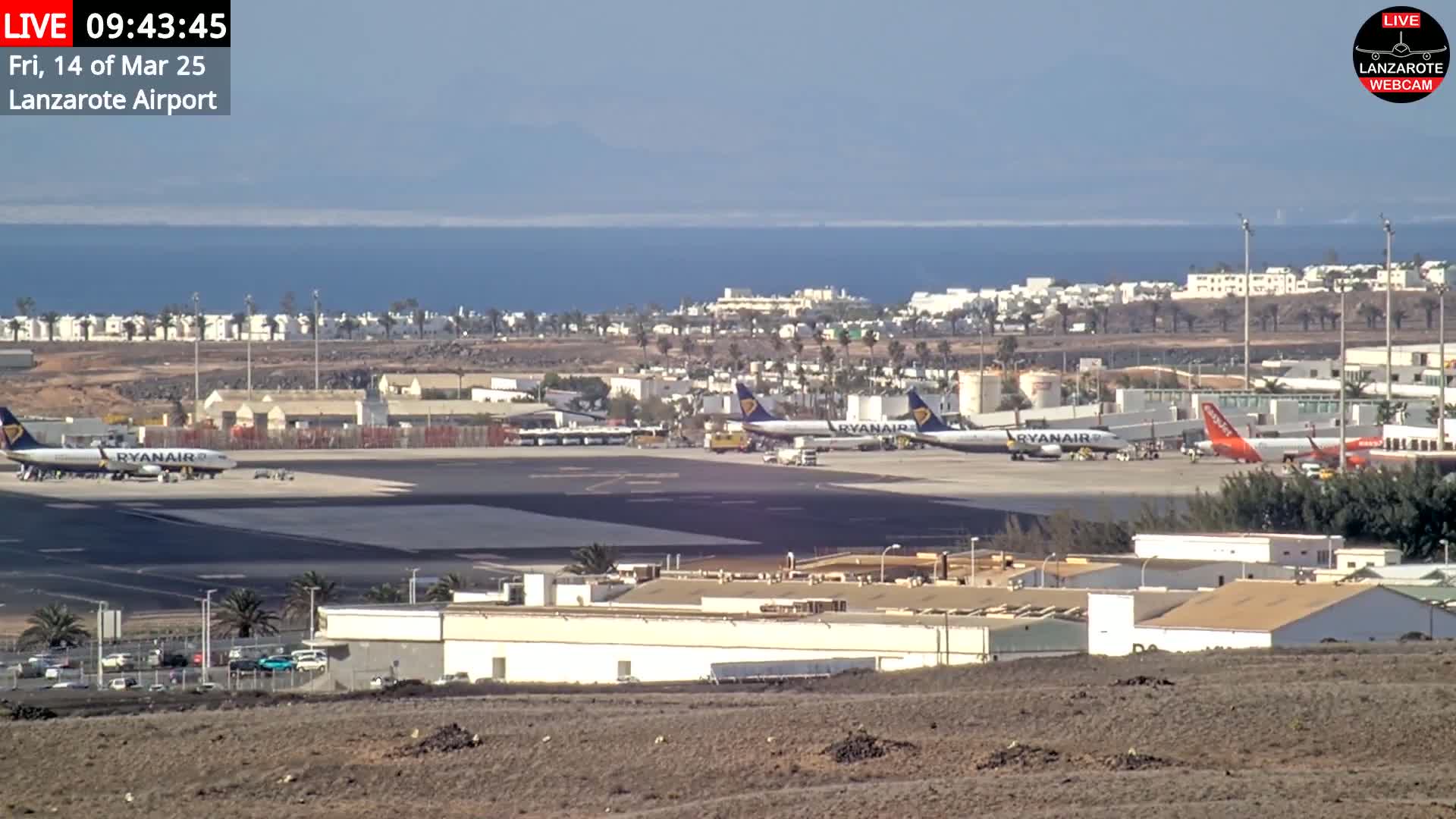 Several airplanes are parked on an airport tarmac near the ocean on a clear day.