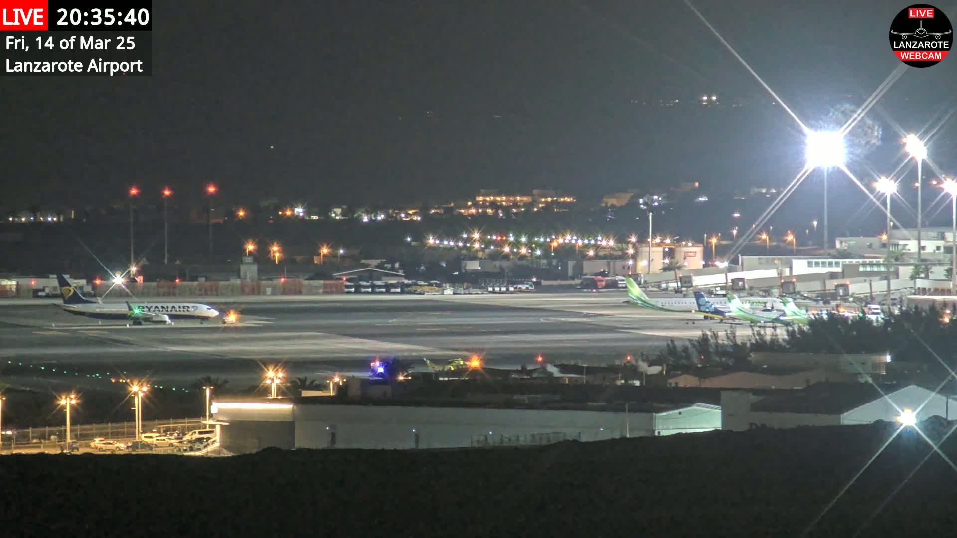 A nighttime view of an airport tarmac with several airplanes, brightly lit buildings, and surrounding area lights.