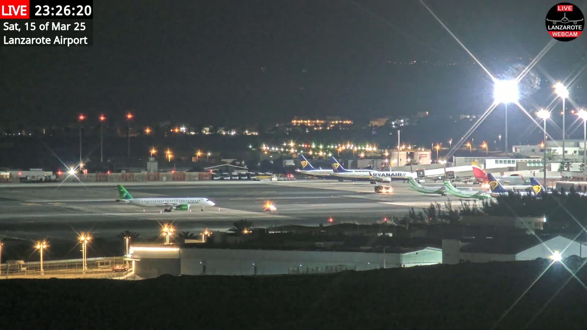 A brightly lit airport tarmac at night shows several airplanes parked, with one taxiing.