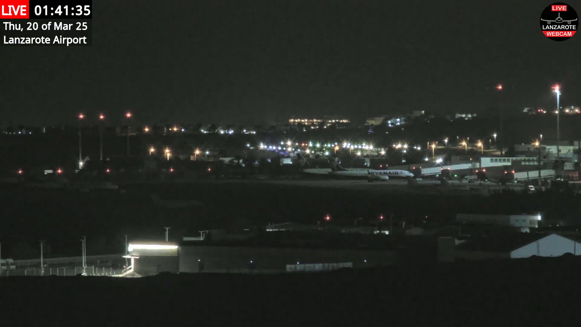 A nighttime view of an airport tarmac with a Ryanair airplane and surrounding buildings illuminated by lights under a clear, dark sky.