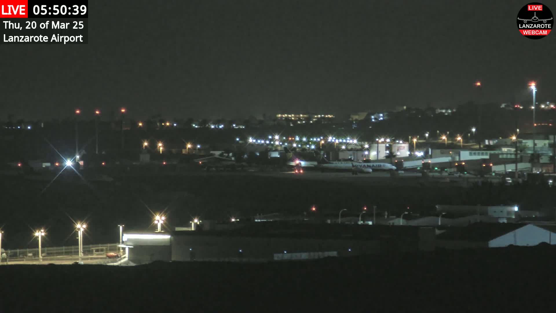 A nighttime long shot of an airport tarmac with several planes and buildings illuminated by lights under a clear, dark sky.