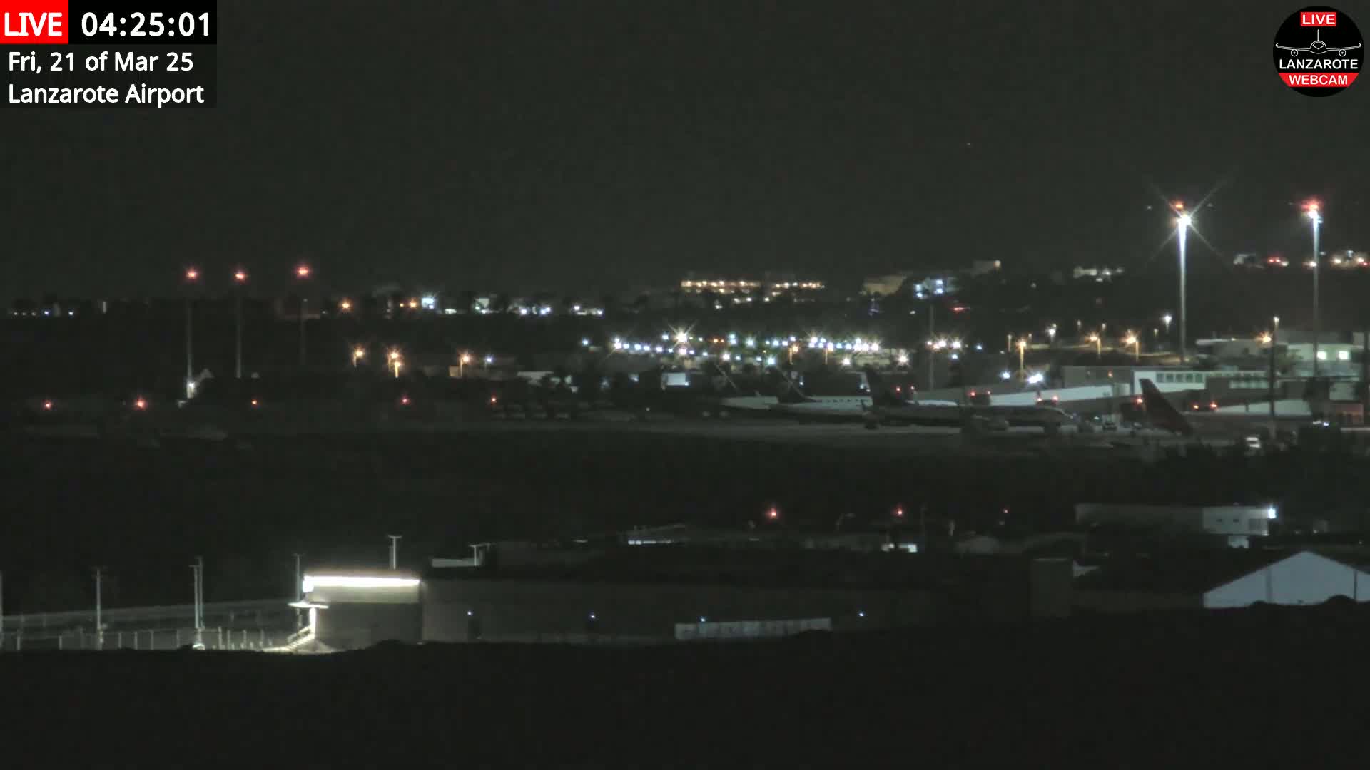 A nighttime view of an airport tarmac with several airplanes, surrounded by buildings and lights under a clear, dark sky.