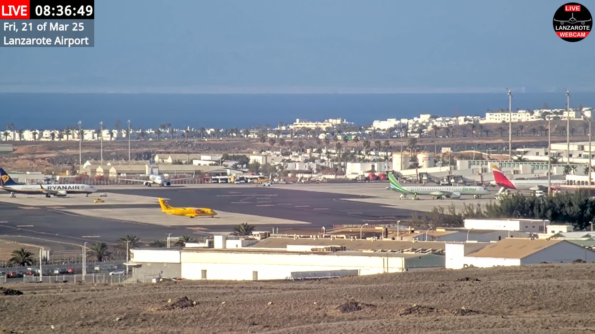A sunny day at an airport shows several airplanes on the tarmac, with a town and ocean visible in the background.