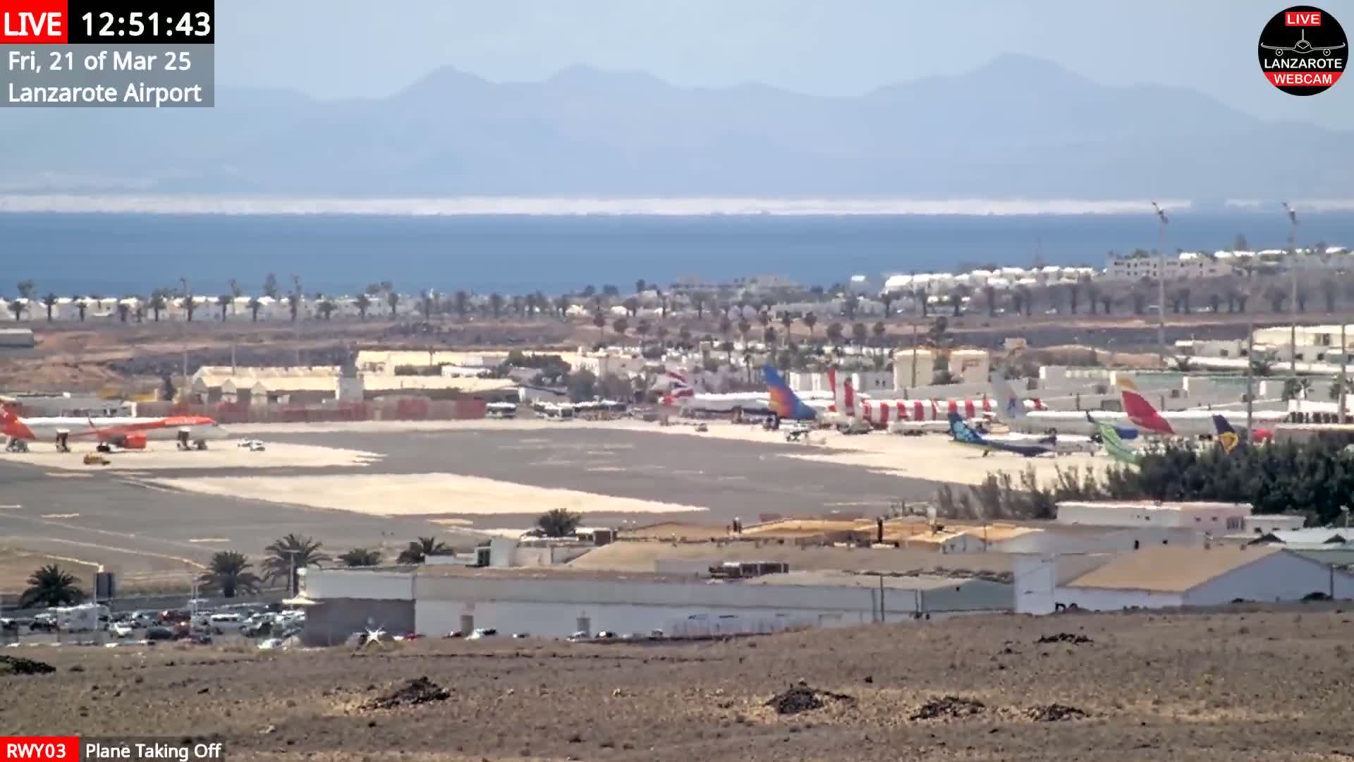 A sunny day at an airport shows several airplanes on the tarmac, some taking off, with a body of water and distant mountains in the background.