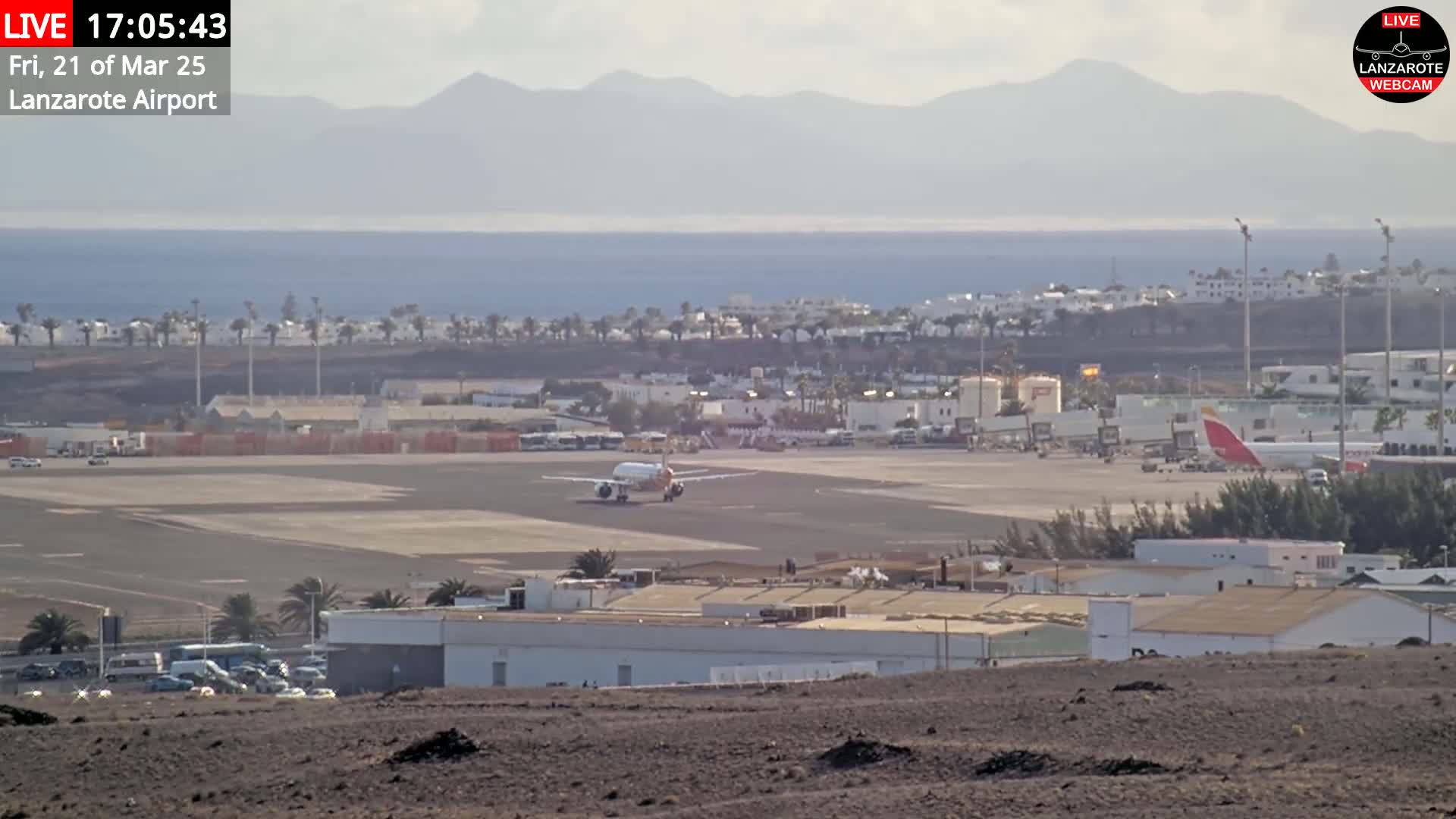 A plane taxis on an airport runway near buildings and a body of water under hazy, sunny conditions with mountains in the distance.