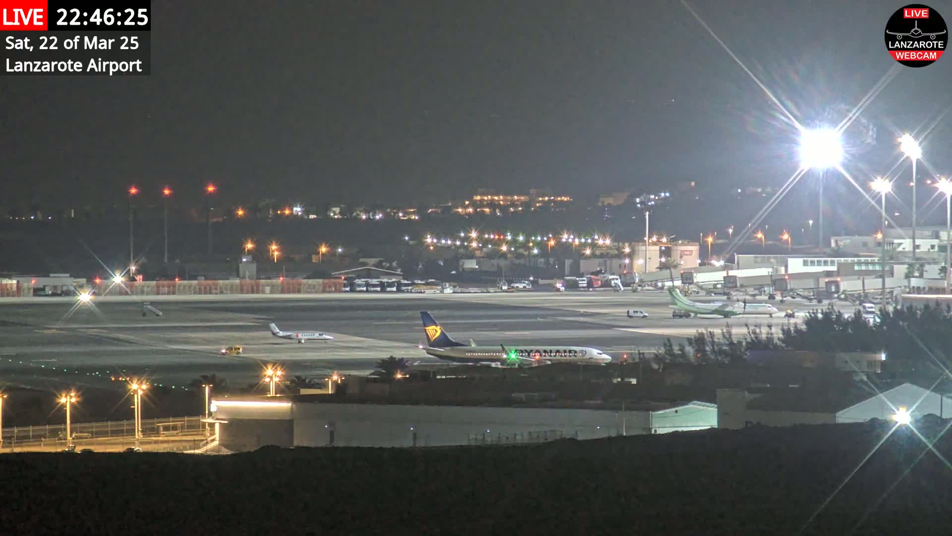 A nighttime view of an airport tarmac with several airplanes, including one with blue and yellow markings, under bright lights.