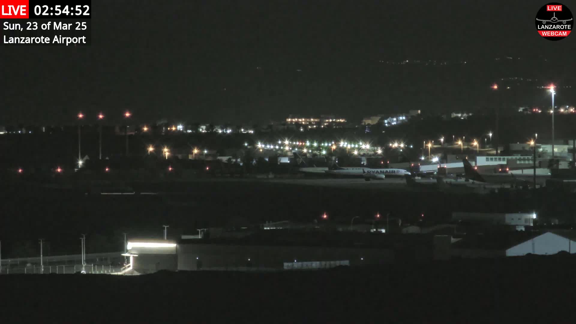 A nighttime long shot of an airport runway with several planes and buildings illuminated by lights.