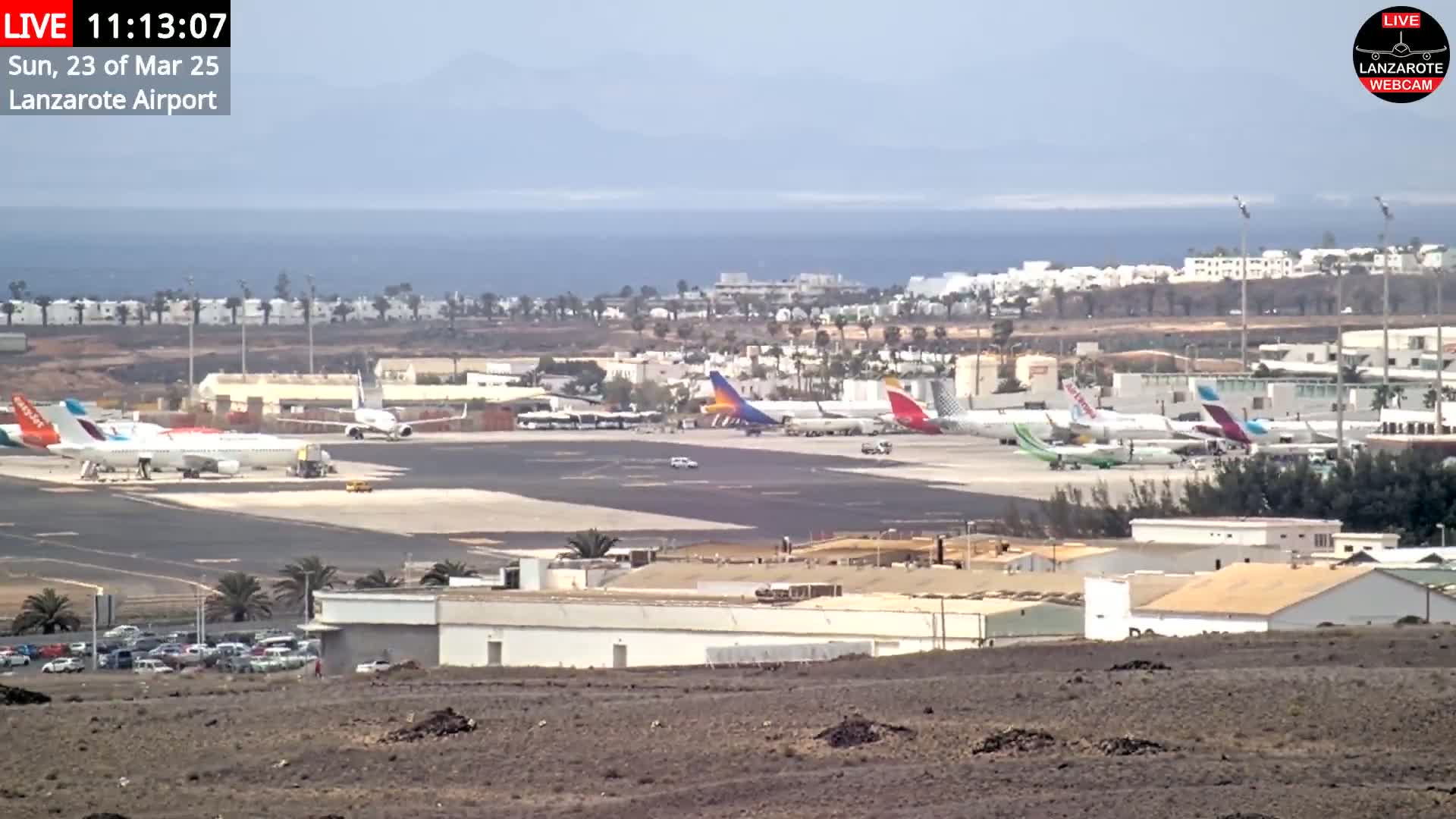 A mostly sunny day at an airport tarmac shows several airplanes parked and a few cars driving on the grounds, with a body of water and distant land visible in the background.
