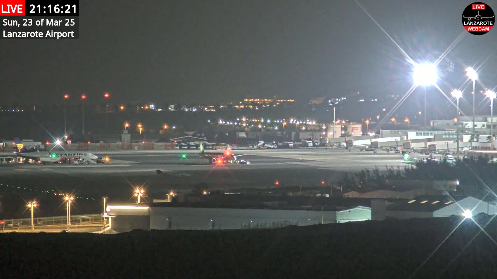 A nighttime view of an airport tarmac with several airplanes, ground vehicles, and buildings illuminated by bright lights.