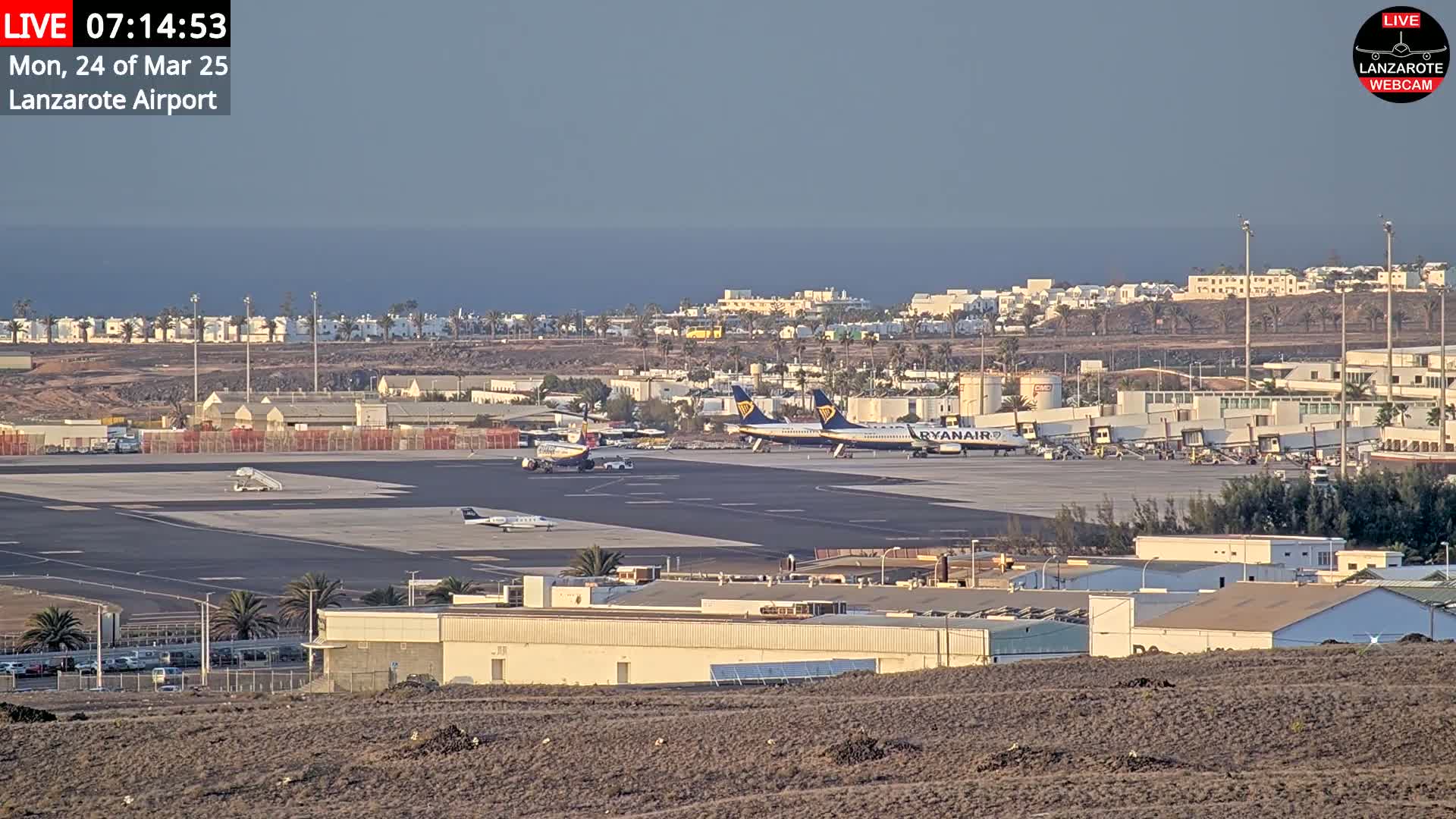 A Lanzarote airport tarmac with several airplanes, including two Ryanair jets, under a hazy sky and overlooking a calm ocean.