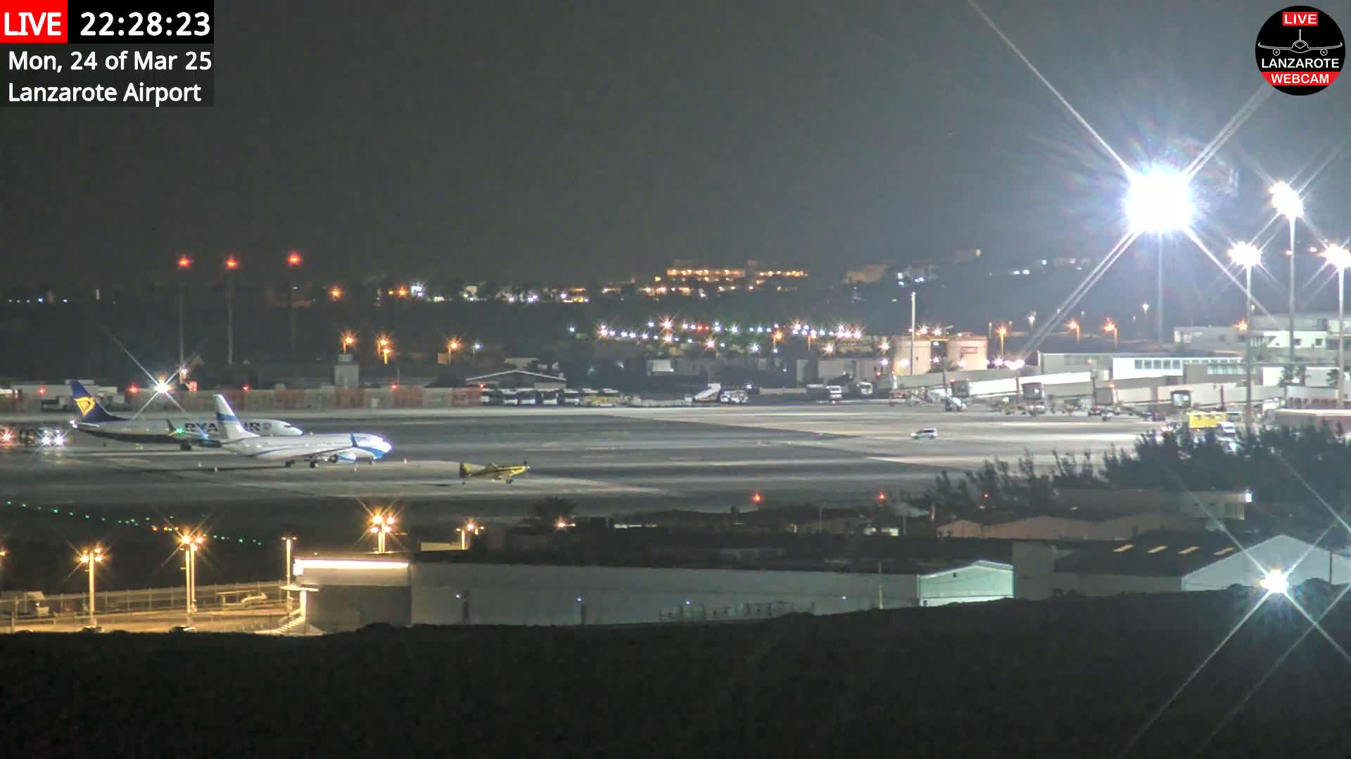 A nighttime view of an airport tarmac with several airplanes, including two parked jets and a smaller plane, under bright lights.