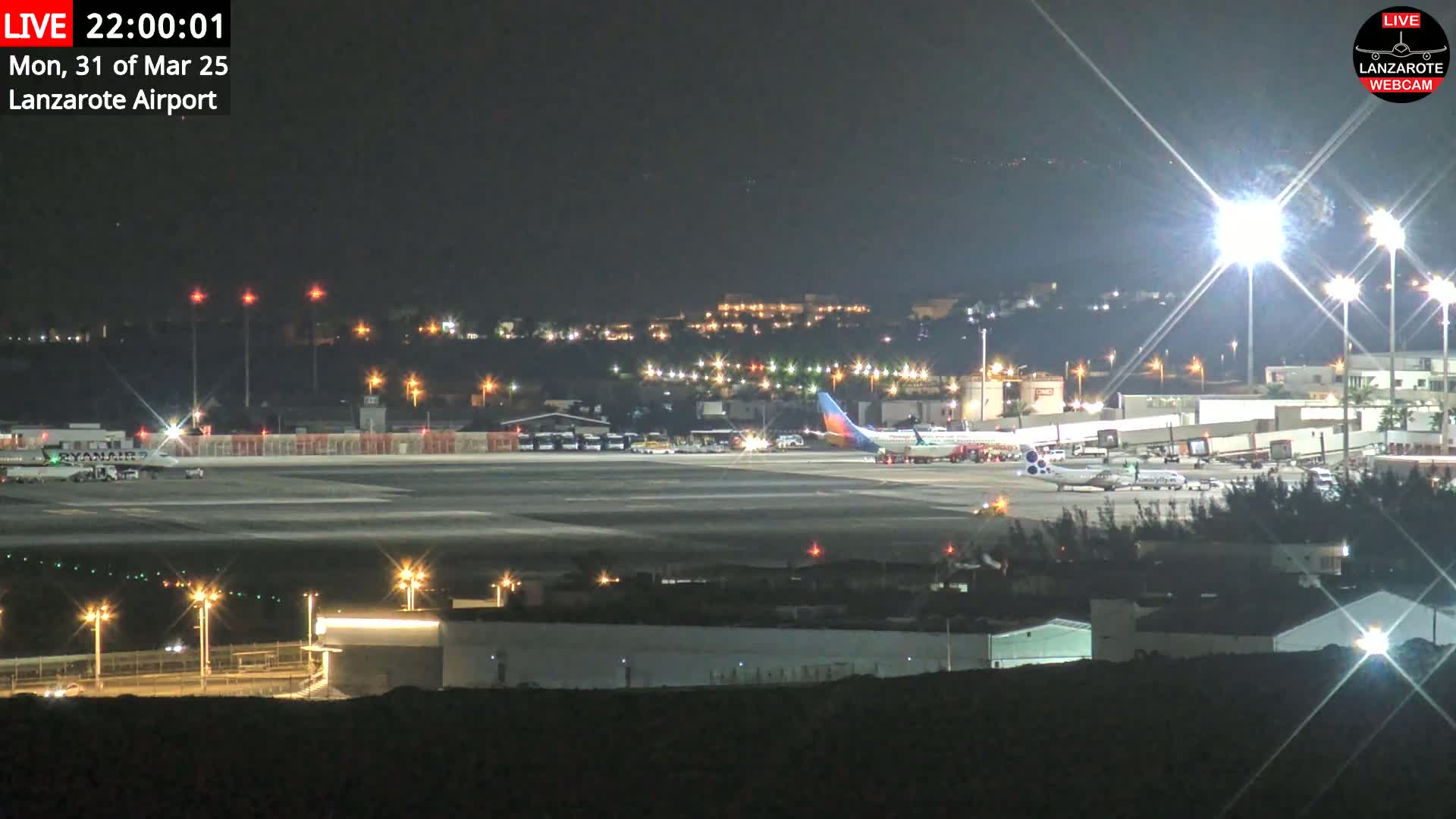 A brightly lit airport tarmac at night shows several airplanes parked near the terminal buildings.