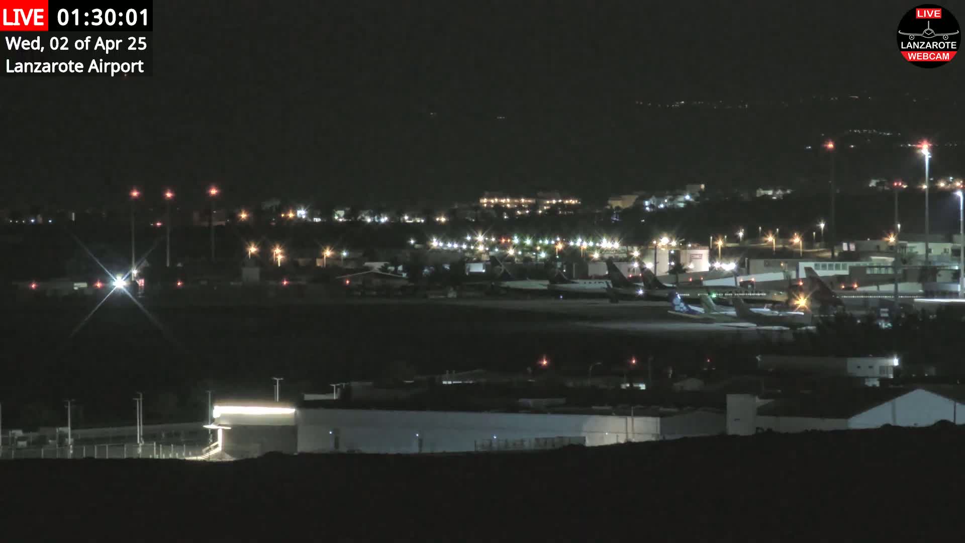 A nighttime view of an airport tarmac with several airplanes parked, surrounded by dimly lit buildings and infrastructure under a clear, dark sky.