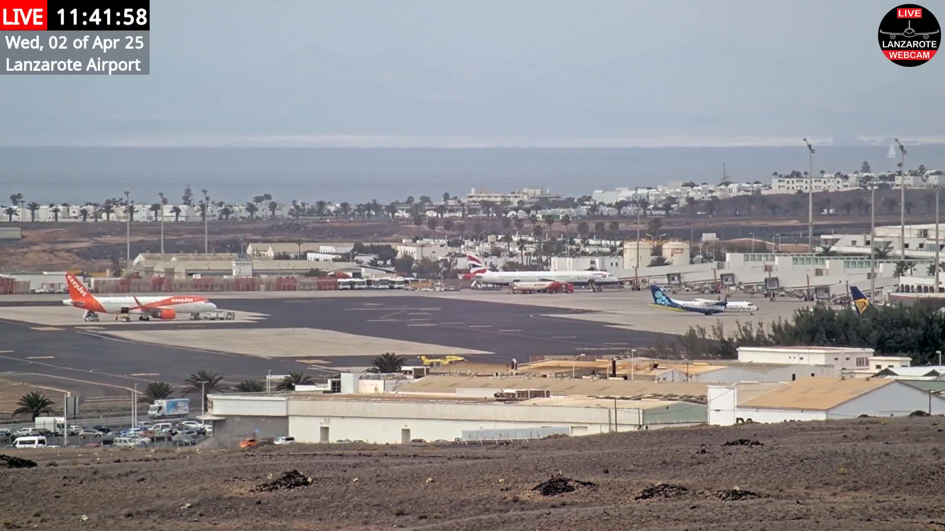 A long shot of an airport tarmac with several planes, including an orange and white plane, under an overcast sky, overlooking the ocean in the distance.