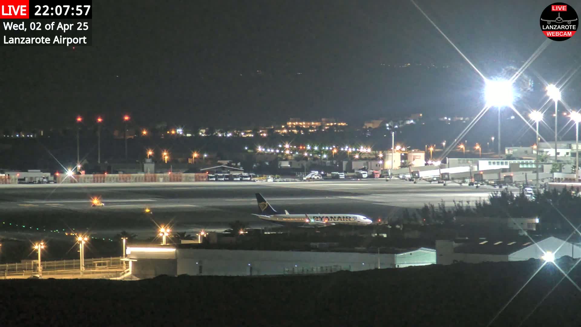 A Ryanair airplane sits on a runway at night, under clear skies, surrounded by airport buildings and illuminated by bright lights.