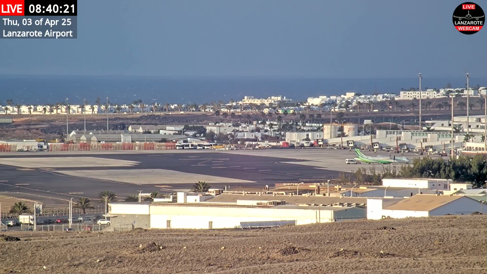 A long shot of an airport tarmac with a plane and surrounding buildings, overlooking a calm ocean under a clear sky.