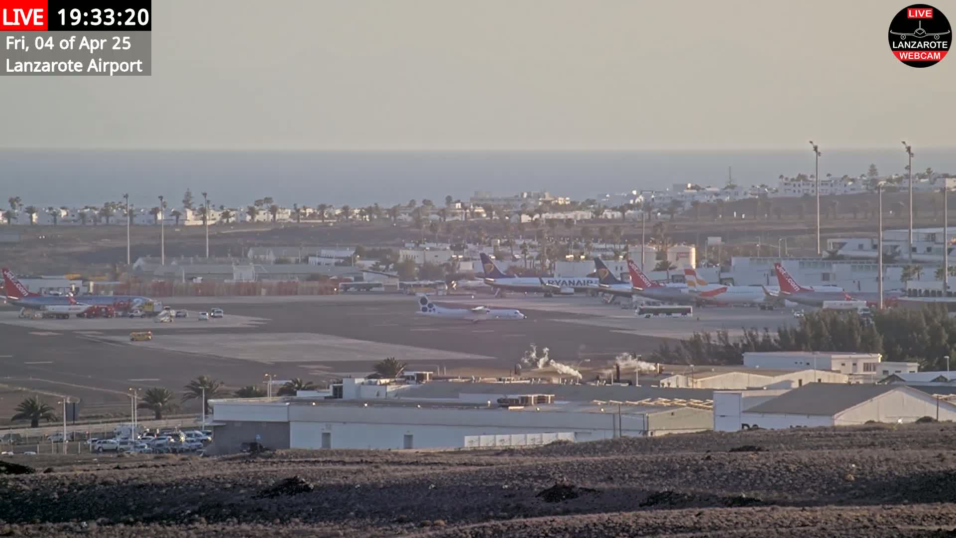 A hazy view of an airport tarmac with several airplanes, ground vehicles, and airport buildings, all situated near a calm ocean under a mostly clear sky.