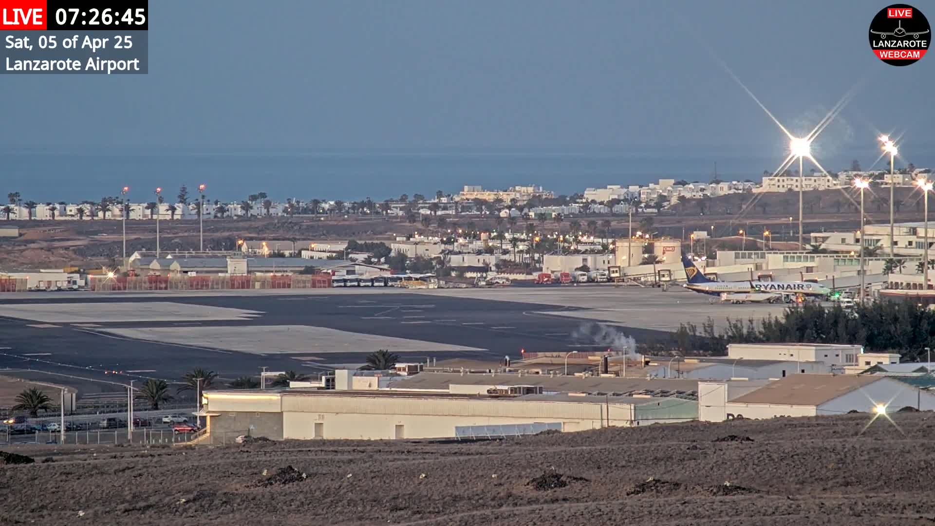 An airport tarmac with a Ryanair plane and surrounding buildings is visible, with a calm ocean and clear twilight sky in the background.