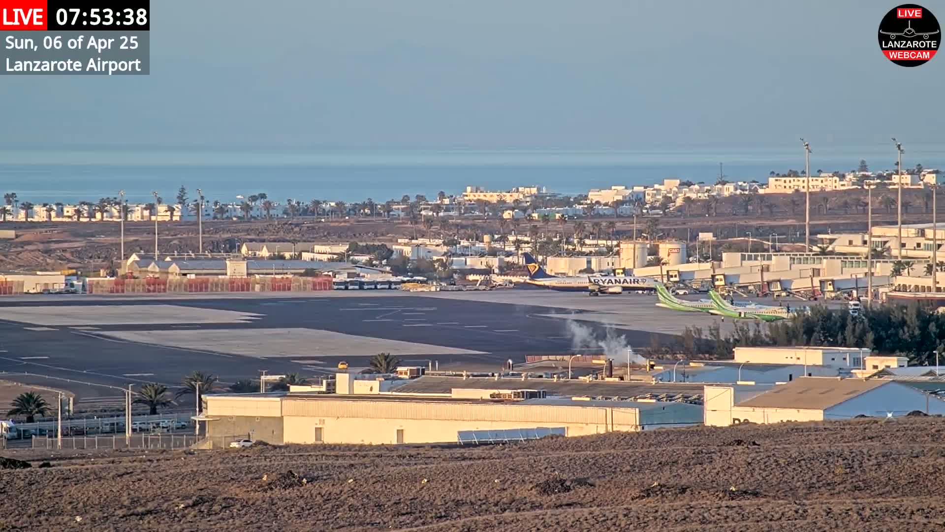 An airport tarmac with several planes and airport buildings is visible, overlooking a calm ocean under a clear sky.