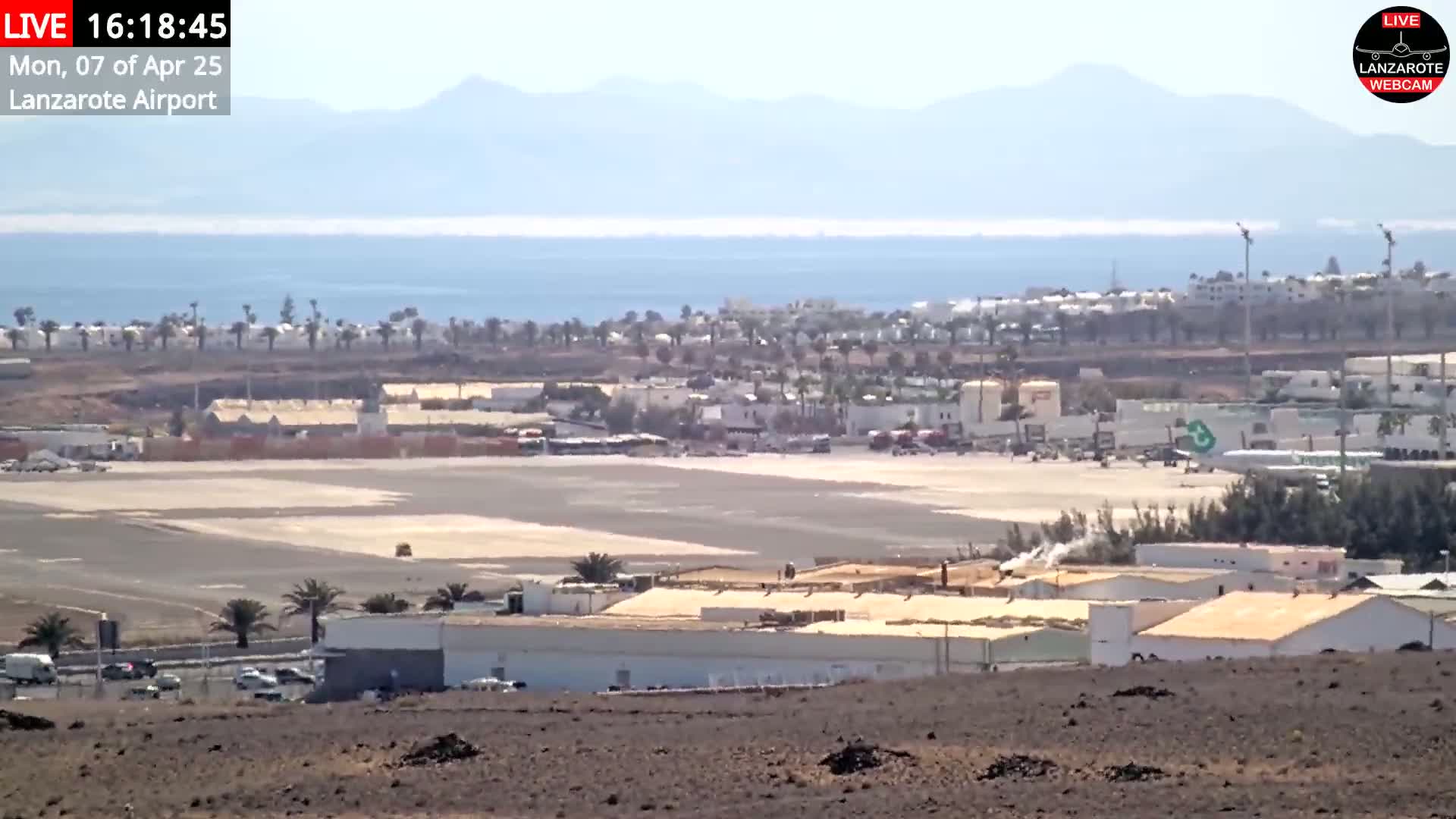 A hazy, sunny day overlooks an airport runway, adjacent buildings, and a coastal town with palm trees, all set against a backdrop of mountains and ocean.
