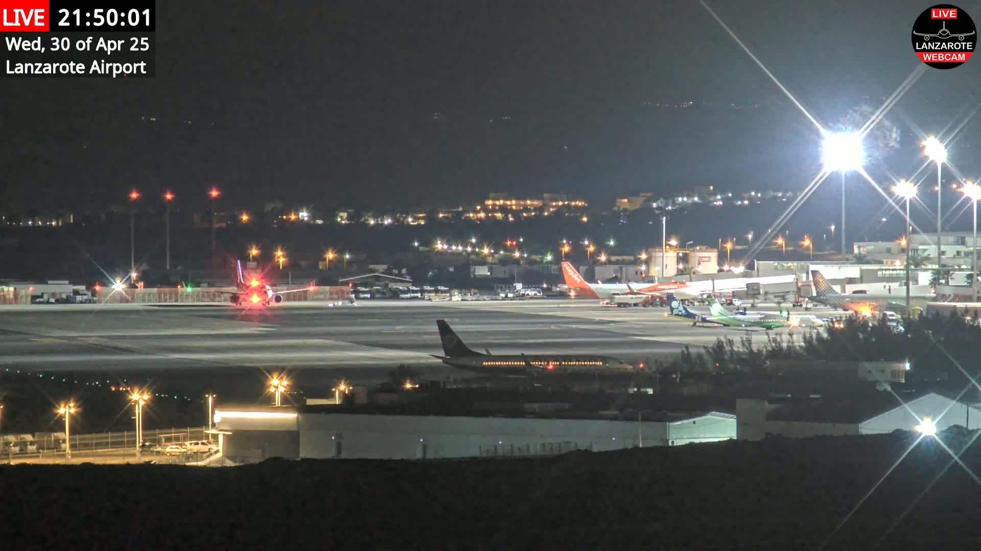 A brightly lit airport tarmac at night shows several airplanes parked and one taxiing, under a clear, dark sky.