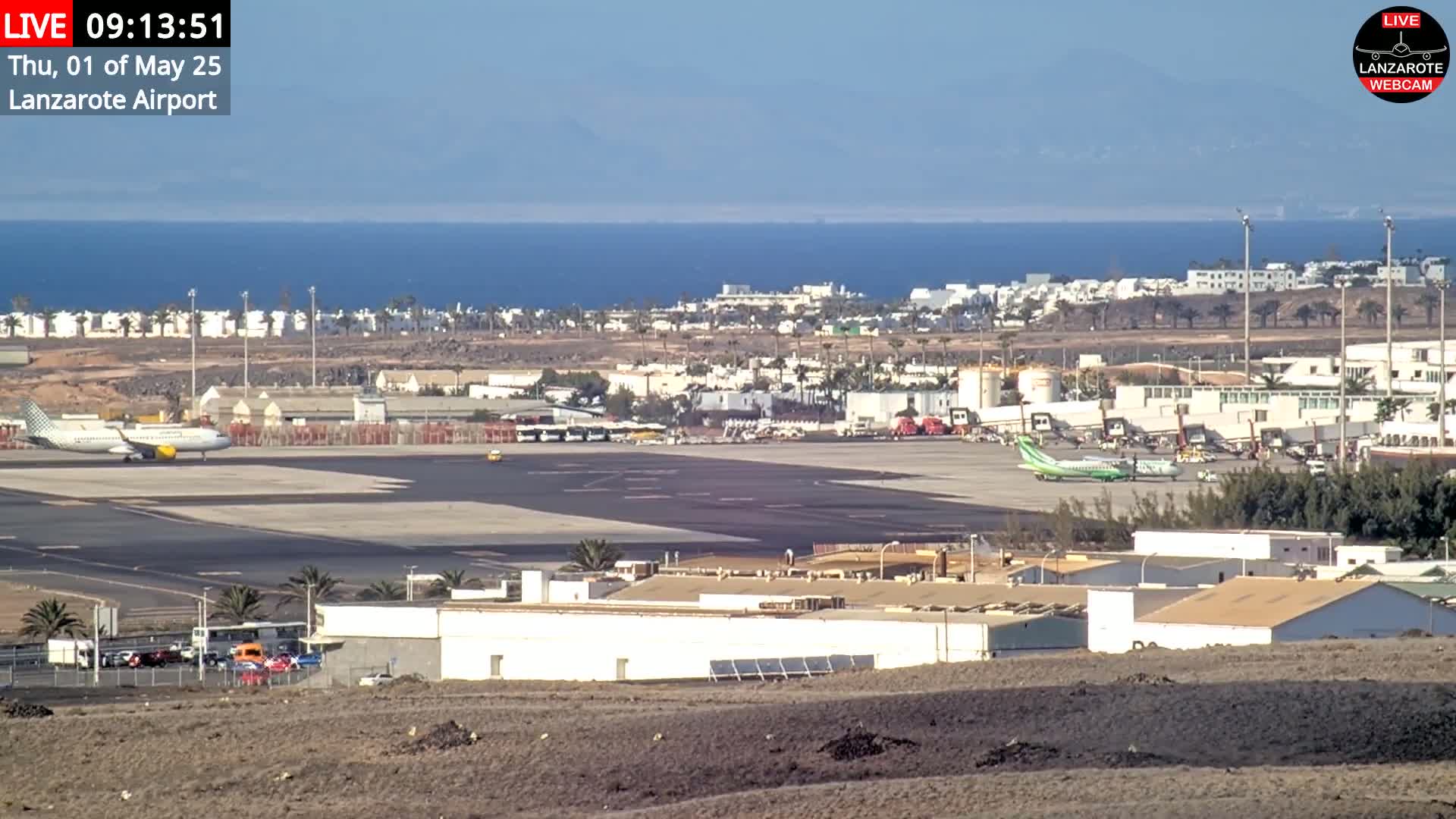 An airport tarmac with several airplanes, airport buildings, and surrounding town under a clear, sunny sky, next to a calm ocean.