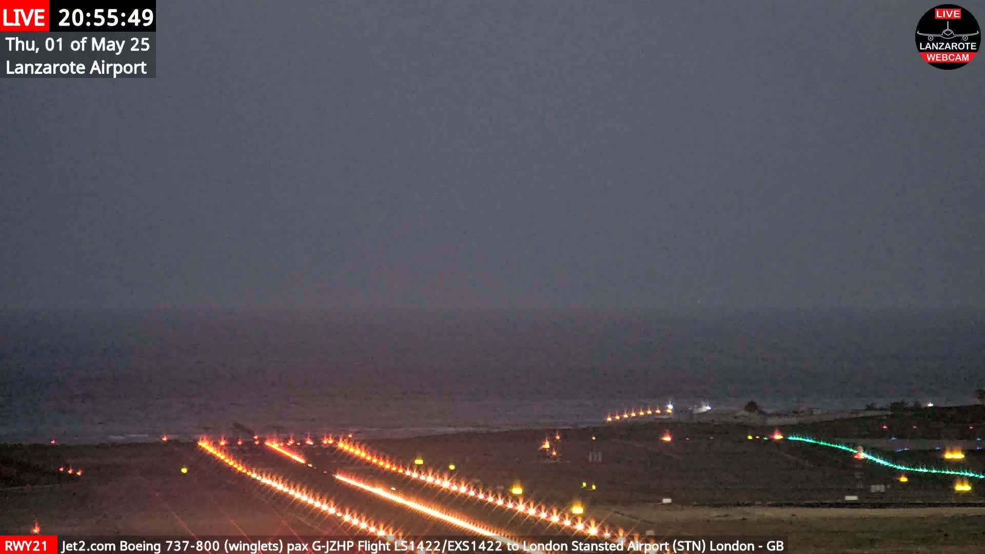 A dark, nighttime view of an airport runway illuminated with lights next to a calm ocean under a dusky sky.
