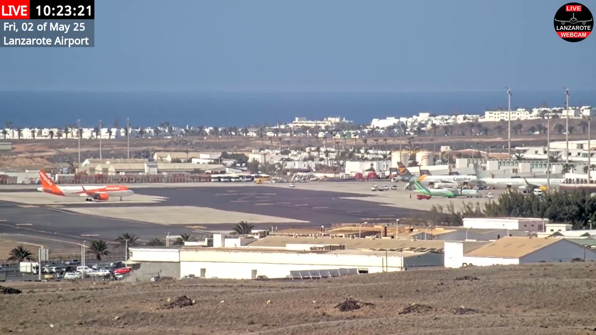 A long shot of an airport tarmac with several planes, including an orange one, under a clear, sunny sky with a calm ocean in the background.