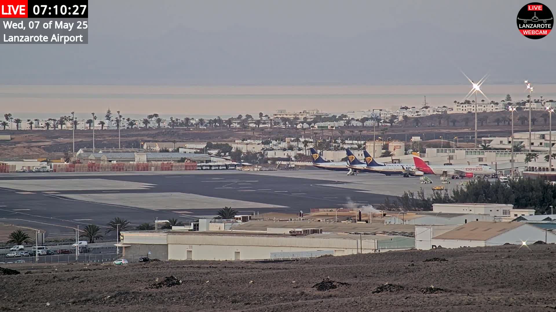 Several airplanes are parked on an airport tarmac near a calm ocean under a hazy sky.