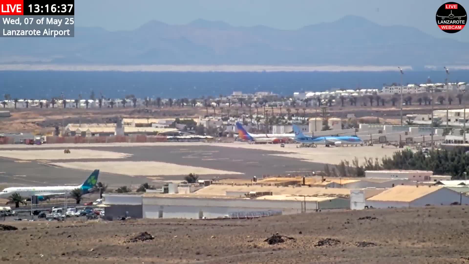 A sunny, long-range view of an airport with several airplanes on the tarmac, adjacent buildings, and a body of water and distant mountains visible in the background.
