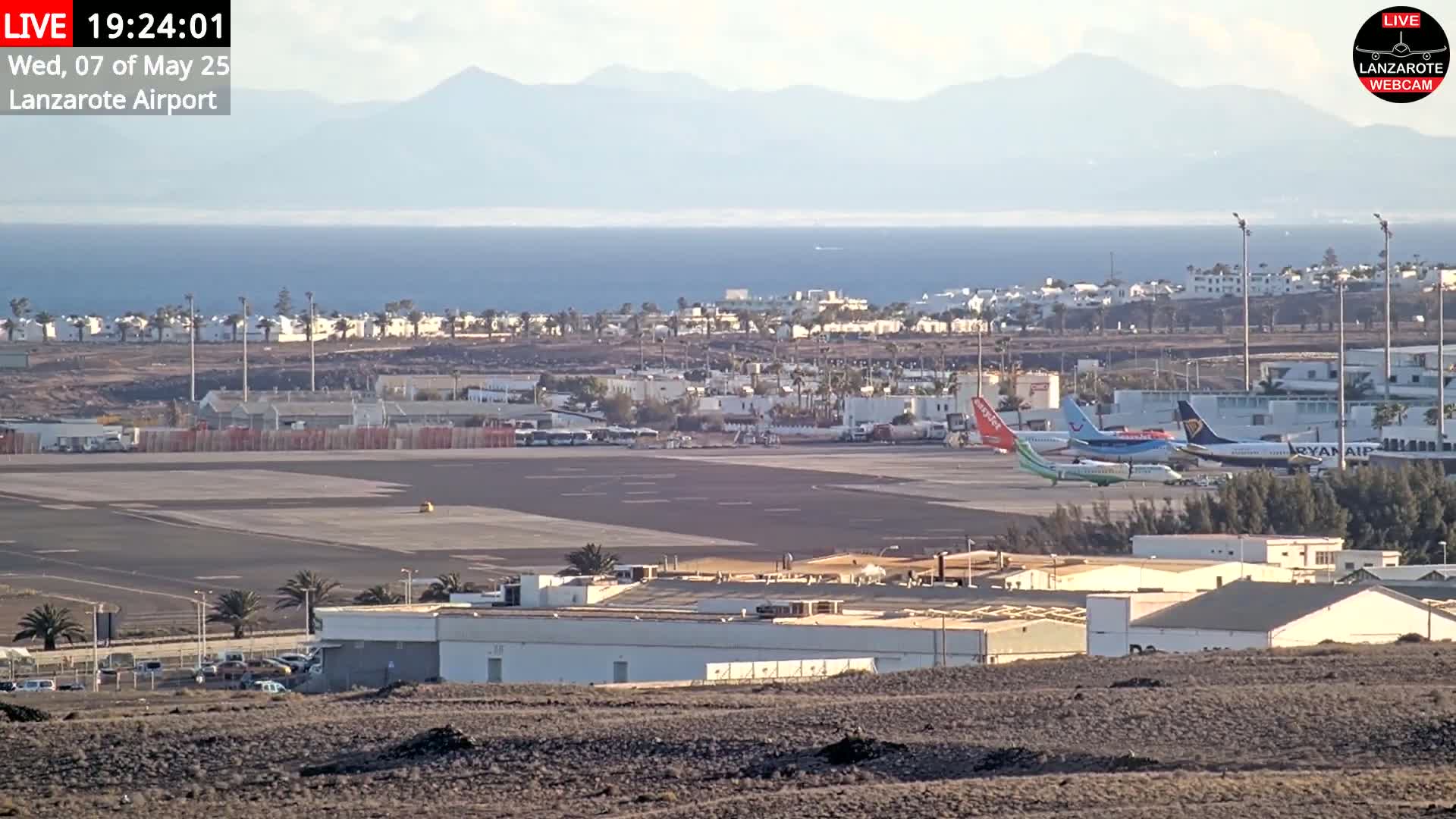 An airport tarmac with several airplanes parked, surrounded by buildings and a town, overlooks the ocean and distant mountains under a mostly clear sky.