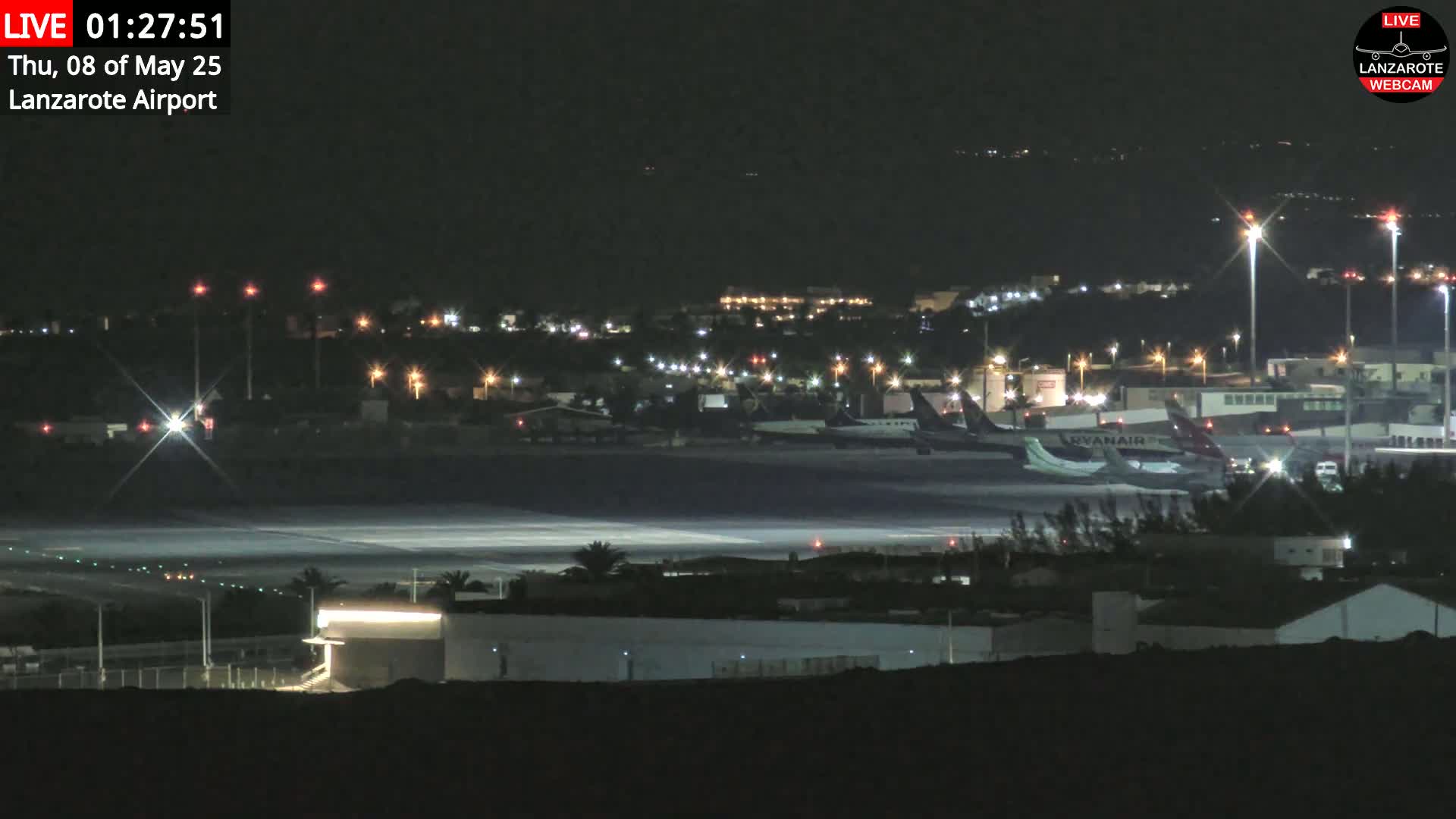 A nighttime view of an airport tarmac with several airplanes parked, surrounded by illuminated buildings and a runway.